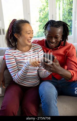 Happy diverse couple using smartphone together, smiling in sunny living room Stock Photo
