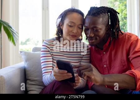 Happy diverse couple using smartphone together, smiling in sunny living room Stock Photo