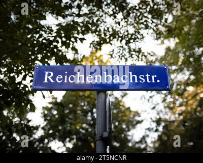 Reichenbachstr. road name sign in Germany close up. The white letters of the street information are on a blue metal plate. Lush foliage is behind. Stock Photo