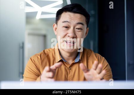 Successful smiling asian businessman talking to colleagues remotely, man using laptop for video call, joyful worker in shirt inside office. Stock Photo