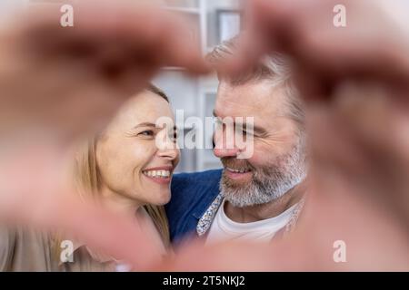 Senior mature couple man and woman showing heart with hands to camera, man and woman in love sitting on sofa in living room, smiling and hugging. Stock Photo