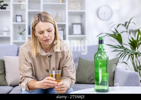 An alcoholic woman drinks strong alcohol, is depressed and sits alone at home on the sofa in the living room. Stock Photo
