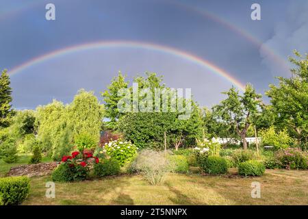 Colorful rainbow in a stormy sky over sunny garden with flowers and fruit trees. Rural landscape. Stock Photo