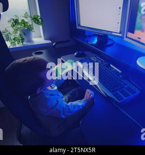 Happy baby is sitting at the computer keyboard with a bottle of milk in his hands. A child with a desktop PC in a home living room drinks milk formula Stock Photo