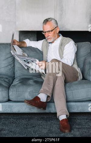 Senior man sitting cozy sofa with crossed leg reading newspaper Stock Photo