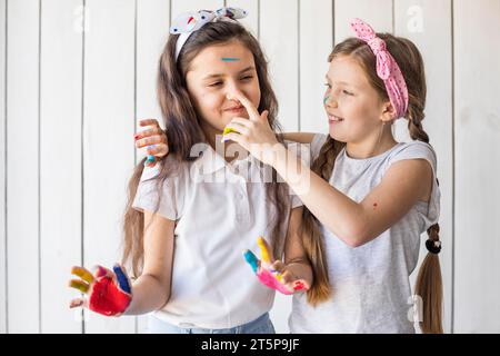 Smiling girl painting her friend s nose with color standing against wooden wall Stock Photo