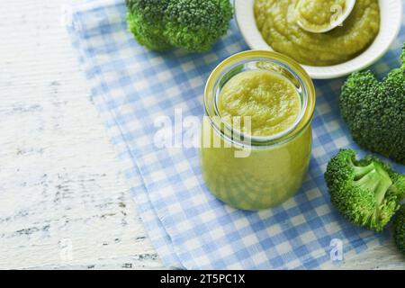 Green broccoli baby food in white bowl and jar on table. Green baby food. Child first feeding concept. Baby Natural Food. Production and menu of baby Stock Photo