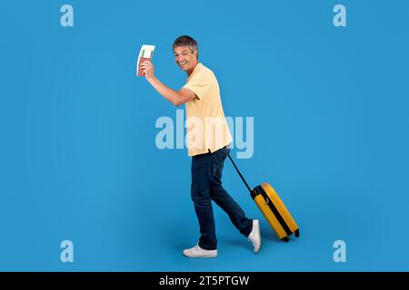 Mature traveler holds boarding pass posing with suitcase, blue background Stock Photo