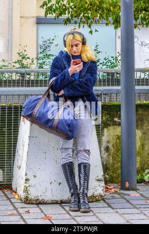 Young woman with holdall, mobile phone and headset, leaning against concrete bollard - Tours, Indre-et-Loire (37), France. Stock Photo