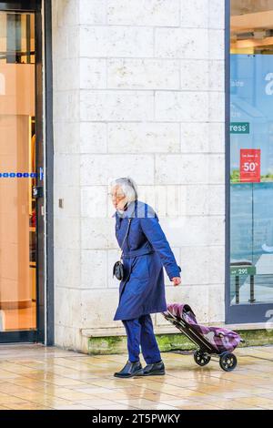 Elderly woman pulling shopping trolley along city pavement in wet weather - Tours, Indre-et-Loire (37), France. Stock Photo