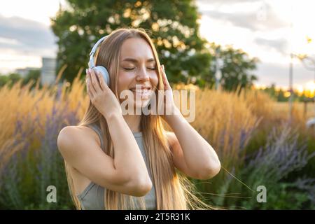 Cheerful young woman enjoys listening to music and singing while sitting in park at sunset. Happy lady in headphones rests spending time in nature Stock Photo