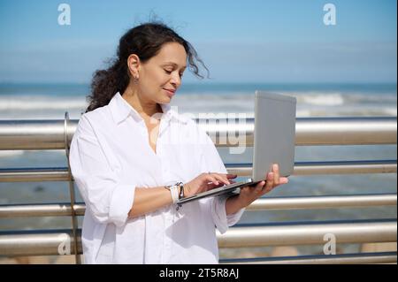 Confident curly haired multi ethnic brunette, business lady in white casual shirt, typing text on laptop while online working outdoors against seashor Stock Photo