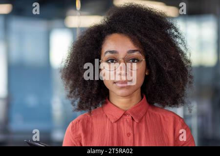 Close up portrait of young beautiful successful business woman, hispanic woman serious confident looking at camera close up, female worker inside office at workplace in red shirt with curly hair. Stock Photo