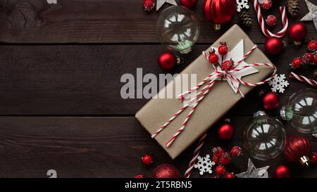 Gift wrapped in craft brown paper lies among holiday decor, red balls, stars, snowflakes and berries on wooden background, copy space, flat lay, top v Stock Photo