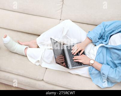 A lady with broken foot  consulting with doctor online using her laptop. Idea of telemedicine for online patient counseling. Woman seeking remote work Stock Photo