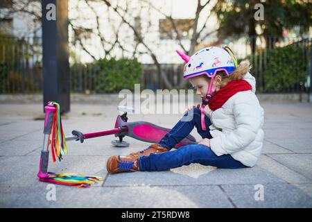 Preschooler girl in unicorn helmet sitting on the ground after she fell while riding her scooter in park on a spring day. Outdoor sport activities and Stock Photo