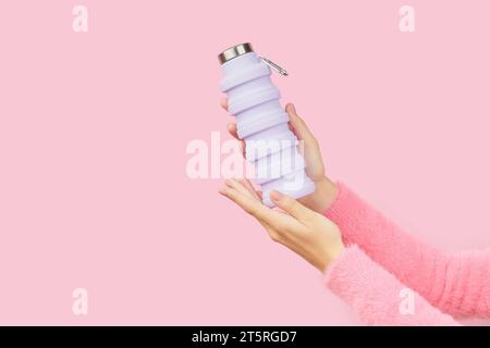 Womans hands holding collapsible reusable lilac water bottle on pink background. Sustainable lifestyle Stock Photo