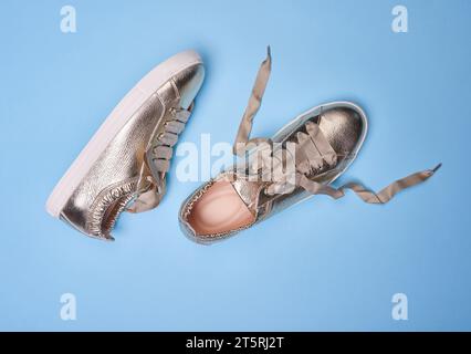 Youth golden metallic sneakers with wrinkled leather details and white rubber soles on a soft blue background. Close-up view. One shoe has untied shoe Stock Photo