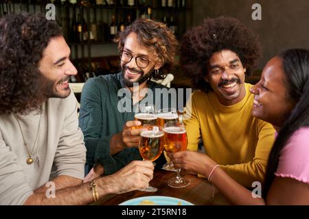 Group of young people having fun and drink beer in a bar toasting. This four multi-ethnic friends Stock Photo