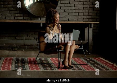 Pretty black woman making a video call on her laptop while sitting in the chair. Joyful girl talking with her friends or family via Internet. Video ca Stock Photo