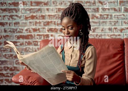 Beautiful black woman with dreadlocks focused on reading a newspaper while sitting on a red leather couch against a red brick wall with copy space.  Y Stock Photo