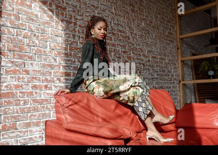 Pretty African girl wearing stylish outfit, sitting on the back of the red leather sofa in the room against a brick wall. Sunbeam lighting falls on pe Stock Photo