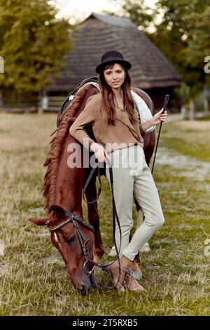 Young woman in black hat stands on grass near a brown horse grazing grass. Horses being fed. Gift certificate for hippotherapy and emotions. Horse in Stock Photo