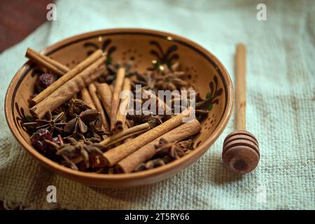 Cinnamon sticks, star anise, and dried rose hips inside a brown bowl with a spoon for honey on the side on linen tablecloth. Preparing Mulled Beverage Stock Photo