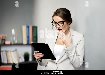 Pretty woman in formal outfit and glasses expressing surprise emotion while using tablet. Reading shocking news or e-mail. Receiving important message Stock Photo