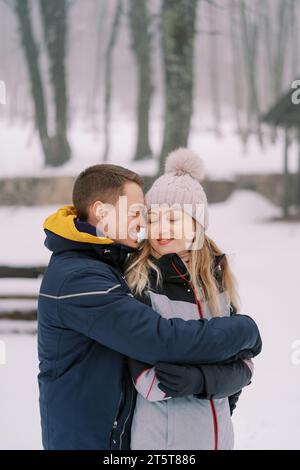 Smiling boyfriend and girlfriend hugging while standing in a snowy park Stock Photo