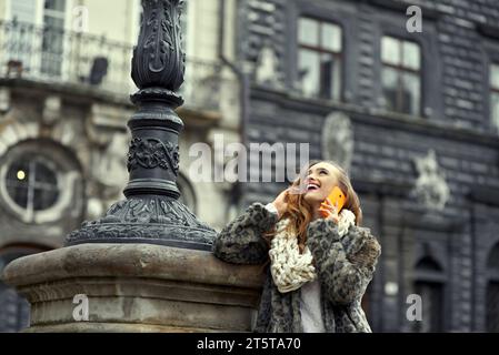 A young woman in a fur coat happily talks to her friend on the phone, telling a funny story. Girl looks up. Talking with relatives about unexpected in Stock Photo