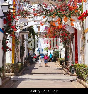 Cascading bougainvillea flowers in vivid red, magenta and orange, frame the streets of Playa de Mogan, Gran Canaria, where a couple of tourists walk, Stock Photo