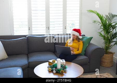 Happy caucasian woman in santa hat using laptop and smiling at christmas at home Stock Photo