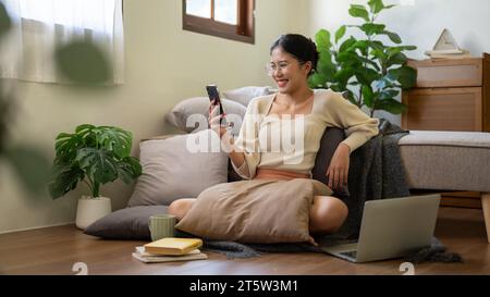A pretty and happy young Asian woman is sitting on the floor beside her bed and using her smartphone, watching videos online or scrolling on social me Stock Photo