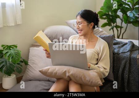 A happy and charming young Asian woman is working from home, researching information in a book, and working on her laptop while sitting in her bedroom Stock Photo