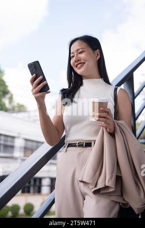 A beautiful Asian businesswoman is using her smartphone, chatting or checking messages while walking down the stairs in the city. Urban city life, peo Stock Photo