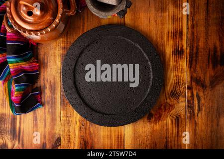 Empty stone comal plate on wooden rustic table, colorful typical mexican fabric with clay pot and stone molcajete, traditional kitchen utensil used in Stock Photo