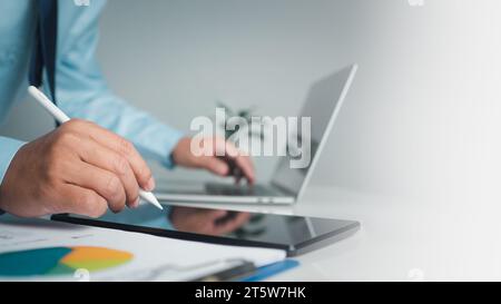 Businessman holding pen pointing at tablet and using computer to work in modern office with copy space. Stock Photo