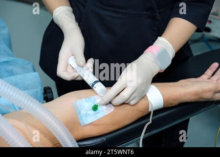 A doctor wearing sterile gloves injects medication intravenously into the patient's arm with a syringe. Injecting anesthesia through a catheter on a person's arm on the operating table. Stock Photo