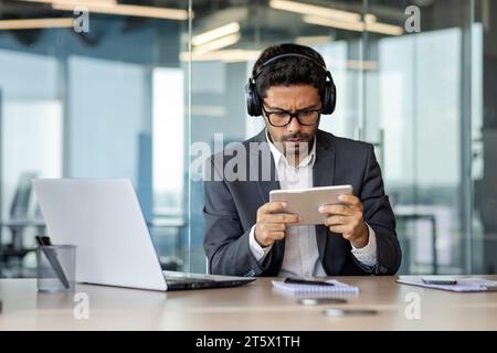Upset and disappointed man in business suit playing game on tablet computer inside office at workplace, losing boss in online games. Stock Photo