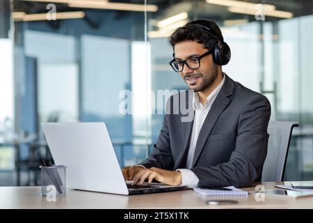 Successful indian programmer working inside office with laptop, man in headphones and business suit, typing code on keyboard, developing new software. Stock Photo
