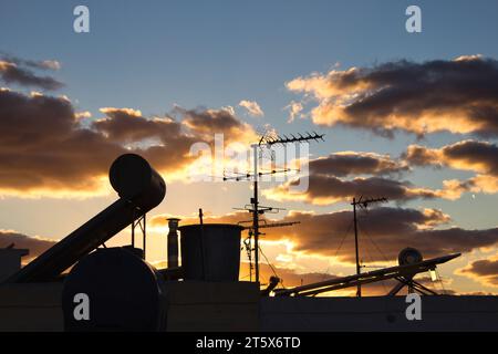 Rooftops silhouetted against a blue cloudy sky at dusk - tv aerials, water tanks and solar panels on rooves - orange sunlight breaking through Stock Photo