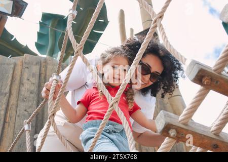 A cheerful mother encouraging her daughter playing together on a rope ladder in a public park - encouragement concept Stock Photo