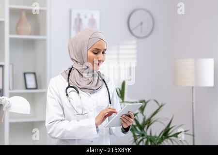 Muslim woman in white medical coat standing inside medical office of clinic, female doctor using tablet computer thinking and concentrating, concentrated woman at work in hospita.l Stock Photo