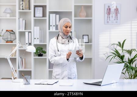 Muslim woman in white medical coat standing inside medical office of clinic, female doctor using tablet computer thinking and concentrating, concentrated woman at work in hospita.l Stock Photo