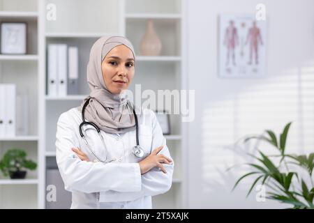 Portrait of serious confident successful female doctor in hijab in white medical coat looking at camera with crossed arms, clinic worker working inside office medical room. Stock Photo