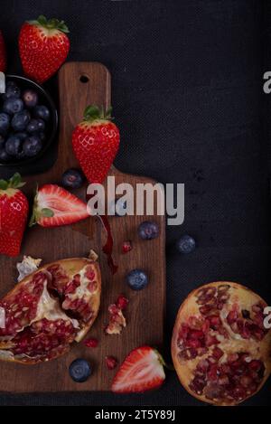 Strawberries, blueberries and pomegranates exposed on a wooden board in dark background. Stock Photo