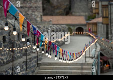 Garland of colorful flags and glowing light bulbs against a defocused background of an stone castle. Stock Photo
