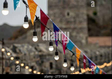 Garland of colorful flags and glowing light bulbs against a defocused background of an stone castle. Stock Photo
