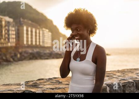 Horizontal photo with copy space of an afro woman smiling while talking to the mobile during sunset in the sea Stock Photo